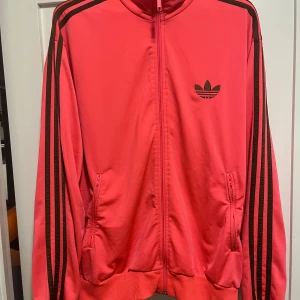 Röd Adidas track jacket med svarta ränder - Säljer en Adidas Originals jacka med svarta klassiska tre ränder ”3 stripes” längs ärmarna och broderad Adidas-logga på bröstet. Jackan har dragkedja framtill, ribbade muddar och två fickor med dragkedja. Perfekt för en sportig och retro vibe. En brutalt snygg Adidasjacka i Korallfärg. Mycket fint skick ! Xl & normal i storlek. Nypris 1299kr