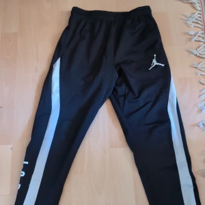 Svarta Jordan träningsbyxor med vit detalj - Snygga svarta träningsbyxor från Jordan med vit logga på låret och breda vita ränder längs sidorna. Byxorna har elastisk midja och texten JORDAN tryckt längs ena benet. Perfekta för sport eller chill, tillverkade i mjukt syntetmaterial.