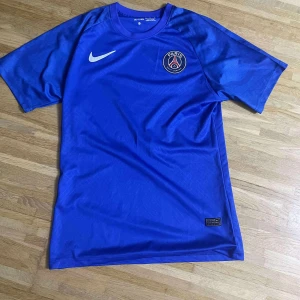 Blå PSG fotbollströja från Nike - Snygg blå fotbollströja från Nike med PSG-logga på bröstet och klassisk vit swoosh. Tröjan är tillverkad i lätt och ventilerande material, perfekt för träning eller match. Rak passform och korta ärmar för en sportig look.