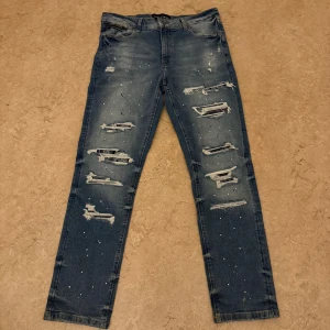 Blå distressed jeans Supply & Demand - Säljer ett par blå jeans från Supply & Demand med slitna detaljer och hål framtill. Jeansen har skinny passform, normal midja och är dekorerade med små vita stänk. Klassisk femficksmodell och stretchigt jeanstyg för skön känsla.