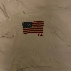 Vit stickad tröja från Ralph Lauren - Vit stickad tröja från Ralph Lauren med amerikansk flagga och RL-broderi på bröstet. Tröjan har rund halsringning, långa ärmar och ribbade muddar. Perfekt för dig som gillar klassisk stil. Skriv ifall ni vill ha fler bilder och mått, vet inte original storleken men skulle säga att den är xxs eller 158-164. Inga defekter 