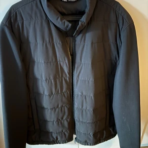 Svart quiltad pufferjacka från Zara - Svart pufferjacka från Zara med quiltad design och hög krage. Jackan har dragkedja framtill och är tillverkad i syntetmaterial. Perfekt för kyligare dagar och har en clean, stilren look.