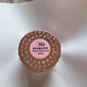 NG Starline Thick White Gel 30ml - NG Starline Thick White Gel i en burk på 30ml. Locket är dekorerat med glittriga stenar och har en rosa etikett. Perfekt för nageldesign och kreativa looks. Produkten är vit och har en tjock gelkonsistens. 25% av 100% 30ml så ca 15ml kvar om jag gissar 