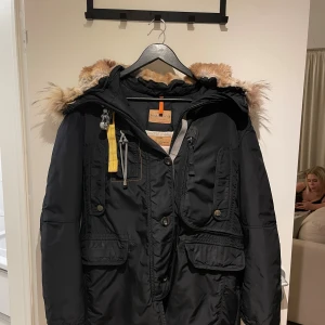 Svart Parajumpers parkajacka med päls - Svart parkajacka från Parajumpers med fluffig beige päls på huvan, coola gula detaljer och flera fickor framtill. Jackan har dragkedja som är sönder men går enkelt att laga är ena dragtappen som är borta, knappar och snörning, samt ribbade muddar vid ärmslut. Perfekt för dig som vill ha en varm och snygg vinterstil.