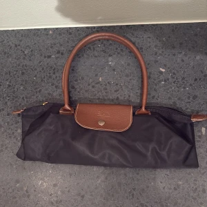 Svart handväska från Longchamp - Snygg svart handväska från Longchamp med bruna skinnhandtag och lock. Väskan har guldfärgad dragkedja med Longchamp-logga och detaljer i brunt skinn. Perfekt storlek för att bära med sig det viktigaste och har en klassisk, stilren look.