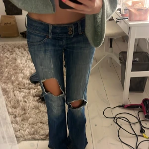 Levis bootcut jeans med slitningar - Säljer ett par blå Levis jeans med bootcut passform och låg midja. Jeansen har slitna detaljer och stora hål på båda benen framtill för en edgy look. Klassiska femficksmodell med dubbla knappar i midjan och snygg tvätt. Perfekta för dig som gillar en avslappnad och trendig stil. Den har ett hål där bak men det syns inte när man har på sig
