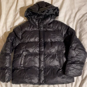Svart pufferjacka med huva - Säljer en svart pufferjacka med huva och dragkedja. Jackan är vadderad och har en klassisk, lite glansig yta. Perfekt för kalla dagar och har en enkel, stilren look som funkar till det mesta.