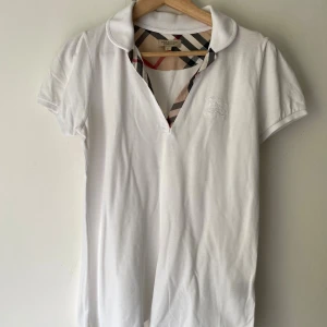 Vintage Vit Burberry blus med klassiskt rutmönster - Säljer en vit kortärmad blus från Burberry London med ikoniskt rutigt mönster på insidan av kragen. Toppen har klassisk krage och v-ringning, tillverkad i mjuk bomull. Perfekt för dig som gillar stilrena och tidlösa plagg med en touch av lyx.