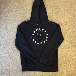 Mörkblå hoodie från Peak Performance - Snygg mörkblå hoodie från Peak Performance med vit logga i cirkel på bröstet. Tröjan har huva med snörning, magficka och långa ärmar. Perfekt för chill eller när du vill vara bekväm men ändå se fräsch ut.
