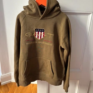 Olivgrön hoodie från GANT med brodyr - Olivgrön hoodie från GANT med broderad logga och amerikanskt inspirerat emblem på bröstet. Klassisk känguruficka, huva och ribbade muddar. Skön och stilren tröja i bomull, perfekt för en avslappnad look.