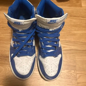 Nike Dunk High blå och vita sneakers - Nike Dunk High sneakers i blått och vitt med klassisk swoosh på sidan. Skorna har snörning, perforerad tå och högt skaft. Tillverkade i skinn med platt sula och rund tå. Perfekt för dig som gillar retrostil och streetwear.