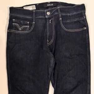 Replay Anbass mörkblå jeans herr - Schyssta mörkblå Replay Anbass jeans med klassisk femficksdesign och kontrastsömmar. Jeansen har raka ben och tydlig logga på bakfickan samt läderpatch i midjan. 