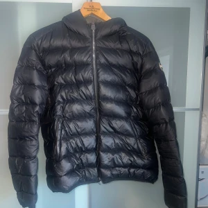 Svart Colmar dunjacka med huva - Svart dunjacka från Colmar Originals med huva och markerad logga på ärmen. Jackan har quiltad design, två sidofickor med dragkedja. Passar en S/M. 