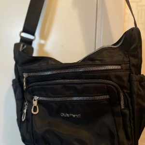 Svart axelväska från Giulia Pieralli - Snygg svart axelväska från Giulia Pieralli med flera praktiska fack och silverfärgade dragkedjor. Väskan har justerbar axelrem med logotyp och är tillverkad i slitstark syntet. Perfekt för dig som vill ha ordning på grejerna och gillar stilren design.