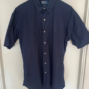 Mörkblå kortärmad skjorta Polo Ralph Lauren - Snygg mörkblå skjorta från Polo Ralph Lauren i 100% bomull. Klassisk krage, korta ärmar och knappar framtill. Diskret broderad logga på bröstet. Perfekt för dig som gillar stilrena och tidlösa plagg med premiumkänsla.