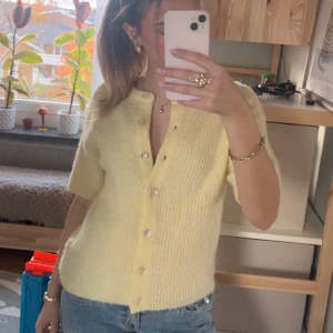 Ljusgul stickad kortärmad kofta - Superfin ljusgul stickad kofta med korta ärmar och pärlemorknappar framtill. Ribbad struktur och rund halsringning. Perfekt att styla med jeans eller kjol för en vardaglig look.