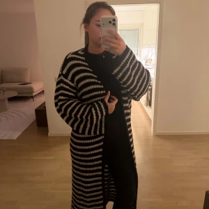 Randig lång kofta i svart och vitt - Lång, oversized kofta med breda svarta och vita ränder. Koftan är stickad med grova maskor och har långa ärmar. Perfekt för lager-på-lager och chill vibes. Ingen knäppning framtill, bara att slänga på sig över en outfit.