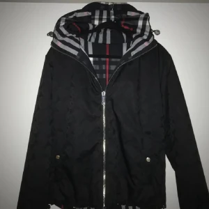 Burberry Reversible Jacket - Snygg vändbar jacka från Burberry med klassiskt rutigt mönster i svart, vitt, grått och rött på ena sidan och helsvart med diskret mönster på andra. Jackan har huva, dragkedja och fickor med tryckknappar. Perfekt för dig som vill ha två stilar i en jacka. 