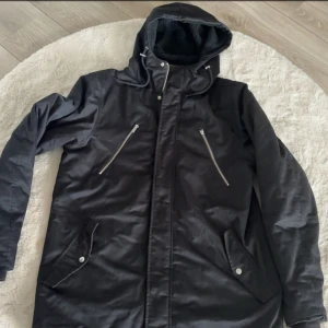 Svart parkajacka med huva och dragkedjor - Svart parkajacka med huva och snörning, två snedställda bröstfickor med dragkedja och två stora fickor med knappar. Fodrad insida och längre modell som passar perfekt när det är kallt ute.