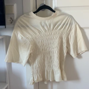 Ljusgul blus med smock från Gina Tricot - Säljer en ljusgul blus från Gina Tricot i storlek S. Toppen har korta ärmar, rund hals och snygg smockad detalj över hela framsidan som ger en cool, rynkad effekt. Materialet är mjukt och känns skönt mot huden. Perfekt för dig som gillar stilrena och trendiga plagg.