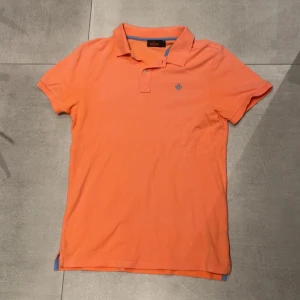 Morris piké  - Snygg orange piké från Morris med klassisk krage och två knappar framtill. Tröjan har korta ärmar och en blå broderad logga på bröstet. Perfekt för en fräsch och fet look under vår och sommar. Men även till fester och kalas under vintern och höst. Tröjan är i storlek M och passar bra på mig som är 185 ungefär, skicket är bra men använt men det syns inte så tydligt!! Hör av dig vid minsta lilla fundering