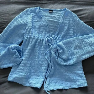 Ljusblå omlottblus med knyt - Supersöt ljusblå blus från gina med omlottknytning i midjan och v-ringning. Blusen har lång ärm och är gjord i ett vågigt, strukturerat tyg som ger en unik look. Perfekt för dig som gillar en luftig och trendig stil.