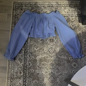 Blå offshoulder blus från Gina Tricot - Säljer en ljusblå offshoulder blus från Gina Tricot. Blusen har långa puffiga ärmar med resår vid ärmslut och en rynkad volangkant upptill. Den är croppad och har en luftig känsla, perfekt för varmare dagar.