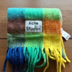 Rutig halsduk Acne Studios i ull - Färgstark och fluffig halsduk från Acne Studios med rutigt mönster i blått, grönt, gult, orange och brunt. Stor logotyp framtill och långa fransar i ändarna. Perfekt statement-piece för att lyfta din outfit under kyliga dagar. Nu med tag. Nypris är runt 3300kr