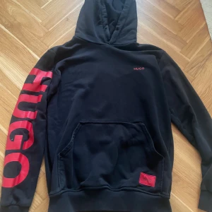 Svart hoodie från Hugo med röd logga - Svart hoodie från Hugo med stor röd logga längs ena ärmen och mindre röd Hugo-text på bröstet. Klassisk känguruficka framtill, huva och ribbade muddar. Snygg kontrast med röda detaljer och perfekt för en streetwear-look.