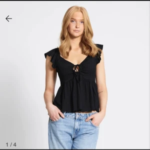 Svart blus med volang och knyt - Supersöt svart blus med volangärmar och v-ringning. Toppen har ett knyt framtill och en lätt rynkad, flowy nederdel som ger en chill och feminin vibe. Perfekt att matcha med jeans eller kjol. Materialet känns mjukt och luftigt.