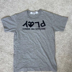 Grå Comme des Garçons Play t-shirt - Snygg grå t-shirt från Comme des Garçons Play med svart tryck framtill och det ikoniska hjärtat med ögon. Klassisk rund halsringning och korta ärmar. Tillverkad i mjuk bomull för en skön känsla. Perfekt för dig som gillar streetwear och stilrena plagg.