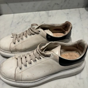 Alexander McQueen vita sneakers - Säljer ett par klassiska vita sneakers från Alexander McQueen med svart detalj på hälen. Skorna har rund tå, platt sula och breda vita snören. Tillverkade i skinn med diskreta hål på sidorna för ventilation. Perfekta för dig som gillar chunky sneakers med stilren vibe. Storlek 45