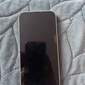 iPhone 13 - Snygg vit iPhone 13 med dubbla kameror och elegant design. Skärmen har en spricka i nedre vänstra hörnet, men övrigt skick ser bra ut. Kamerorna är dekorerade med stenar. Perfekt för dig som vill ha en modern iPhone till ett bra pris.