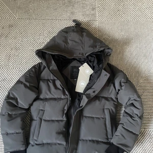 Svart pufferjacka från Canada Goose XS - Säljer en svart pufferjacka från Canada Goose i storlek XS. Jackan har huva, diskret logga på ärmen och klassisk quiltad design. Perfekt för kalla dagar med sin tjocka vaddering och stilrena look. Dragkedja framtill och praktiska fickor.