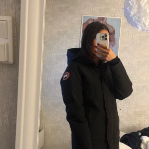 Svart parkas från Canada Goose XS - Säljer en svart Canada Goose parkas i storlek XS med huva och klassisk logga på ärmen. Jackan har dragkedja framtill, två sidofickor och en stilren, lång siluett som håller dig varm. Perfekt för kalla dagar och har en snygg, enkel look.  Pris kan diskuteras 