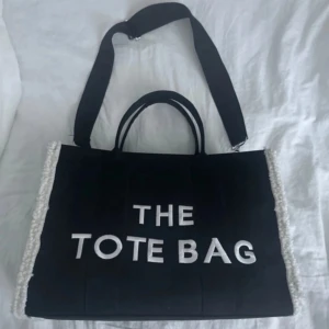 Svart tote bag med vit text - Stor svart tote bag med bred axelrem och två handtag. Väskan har vit broderad text 'THE TOTE BAG' på framsidan och dragkedja upptill. Sidorna har ljusa detaljer i tyg. Perfekt för att bära med sig allt man behöver.