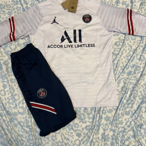 PSG långärmad fotbollströja Jordan - Snygg långärmad PSG fotbollströja i vitt med diskreta ränder, röda och blå detaljer på ärmarna och broderad PSG-logga. Tröjan har dragkedja vid halsen och är tillverkad i lätt, tekniskt material (Dri-FIT ADV). Matchande marinblå byxor med logga och ränder ingår.