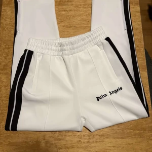 Palm Angels vita mjukisbyxor med svarta ränder - Palm Angels vita mjukisbyxor med svarta sidoremsor och broderad logga på benet. Byxorna har elastisk midja med snörning, raka ben med dragkedja vid benslut och två fickor fram. Snygg sportig look med kontrastdetaljer.