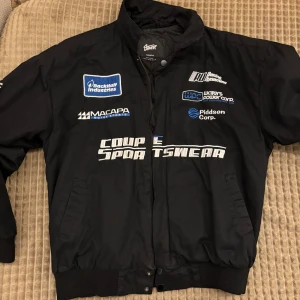 Svart quiltad bomberjacka med brodyr - Svart bomberjacka från SWTZ med broderade racing-inspirerade patches och loggor på bröstet. Jackan har quiltat foder, muddar vid ärmslut och midja samt dragkedja och tryckknappar framtill. Snygg sportig look med tydliga motorsport-vibbar.