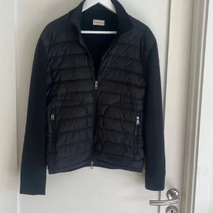 Svart cardigan från Moncler (KVITTO FINNS) - Svart cardigan från Moncler med quiltad front och släta ärmar. Jackan har två dragkedjefickor, hög krage och klassisk Moncler-logga på ärmen. Materialmix med dunfyllning och stickade detaljer ger en clean och sportig look.
