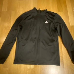 Svart träningskofta från Adidas - Svart långärmad träningskofta från Adidas med hel dragkedja framtill och vit Adidas-logga på bröstet samt stor logga på ryggen. Jackan är tillverkad i polyester och har en sportig, normal passform. Perfekt för träning eller chill.