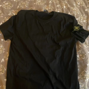 Svart Stone Island t-shirt med patch - Svart t-shirt från Stone Island med klassisk rund hals och korta ärmar. Ikonisk Stone Island-patch sitter fast på vänster ärm. T-shirten är i mjuk bomull och har en clean, stilren look som passar perfekt till streetwear.