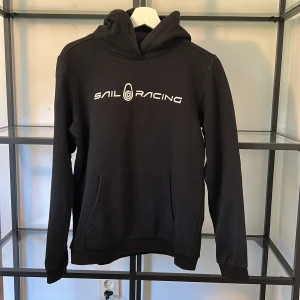 Svart hoodie från Sail Racing - Svart hoodie från Sail Racing med vit logga framtill och liten logga på huvan. Klassisk känguruficka och ribbade muddar. Perfekt för dig som gillar en stilren och sportig look. Nypris 600 kr.