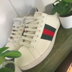 Gucci vita sneakers med röd/grön rand - Säljer ett par klassiska vita sneakers från Gucci i skinn med den ikoniska gröna och röda randen på sidan. Skorna har snörning, rund tå och detaljer i mörkgrönt på hälen. Snygg och tidlös modell som lyfter vilken outfit som helst.