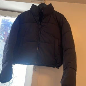 Svart croppad pufferjacka - Svart pufferjacka med croppad passform och hög krage. Jackan har en rejäl dragkedja framtill och två sidofickor. Perfekt för kalla dagar när du vill ha en stilren och trendig look.