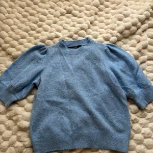 Ljusblå stickad tröja från Vero Moda - Superfin ljusblå stickad tröja från Vero Moda med korta puffärmar och ribbade muddar. Tröjan har en klassisk rund halsringning och är perfekt för lager-på-lager-stil. Mjuk och skön, passar perfekt till jeans eller kjol.