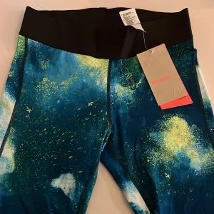 Färgglada tights från H&M Sport - Snygga träningstights från H&M Sport med blått, grönt och vitt mönster och bred svart midja. Perfekta för träning och har en riktigt cool färgexplosion som sticker ut. Tillverkade i stretchigt syntetmaterial för bästa komfort.