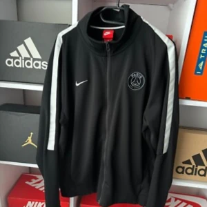 Svart PSG Nike track jacket M - Svart track jacket från Nike med Paris Saint-Germain-logga på bröstet. Jackan har vita ränder längs ärmarna, dragkedja framtill och ståkrage. Perfekt för dig som gillar sportig stil och vill representera PSG.