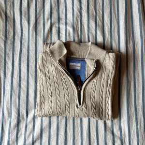 Beige kabelstickad tröja Lindbergh - Säljer en beige kabelstickad tröja från Lindbergh med half zip och ribbade muddar. Tröjan har hög krage och är långärmad, perfekt för lager-på-lager under höst och vinter. Snygg och klassisk stil som funkar till det mesta.