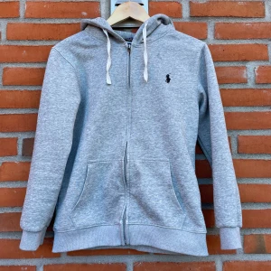 Grå zip up hoodie från Polo Ralph Lauren - Snygg grå hoodie från Polo Ralph Lauren med dragkedja, huva med vita snören och klassisk broderad logga på bröstet. Tröjan har två fickor framtill och ribbade muddar. Perfekt för chill dagar och enkel att matcha med jeans eller joggers. Axelbredd 42 och längd 55-57